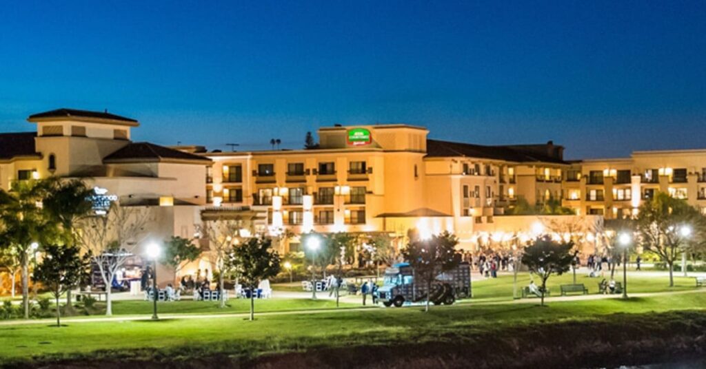 A well-lit hotel with multiple floors and balconies is seen at dusk, surrounded by green lawns, trees, and outdoor seating. People are gathered on the grass as lights illuminate the scene, creating a perfect setting for Deeper Learning in 2026 under a deep blue sky.