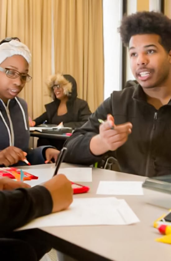 Three people sit at a table in a classroom, engaged in conversation and working on papers, embodying Deeper Learning. In the background, someone smiles in a hooded jacket. The collaborative atmosphere supports focused education and growth.