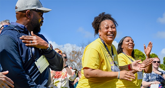 A group of people outdoors, smiling and clapping. Two women in yellow shirts, appearing joyful, stand at the center—showcasing the spirit of Deeper Learning 2026. A man in a dark cap and sunglasses is on the left, with others visible in the background.