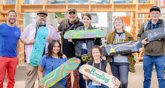 A diverse group of eight smiling people pose indoors, each holding a uniquely painted skateboard deck, highlighting creativity and Deeper Learning in education, standing in front of wooden shelves and bright windows.