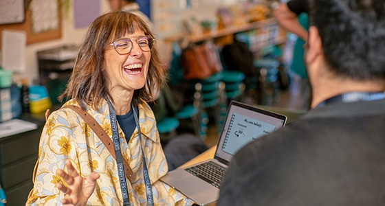 A woman with glasses and a floral scarf smiles and laughs while talking to a man in an indoor education setting, with an open laptop between them and a blurred classroom background—capturing the spirit of Deeper Learning.