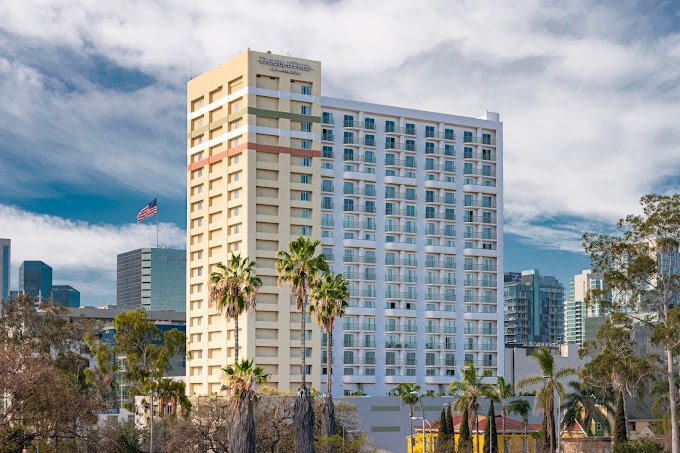 A tall, modern hotel building with balconies stands among palm trees and other skyscrapers in an urban area, under a partly cloudy sky, with the American flag and a Deeper Learning 2026 banner visible in the background.