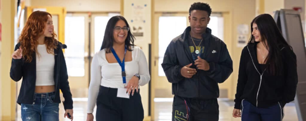 Four young adults walk together down a hallway, smiling and talking after the Grade 9 Summer Symposium. Two women on the left wear casual outfits, while the woman in the center wears glasses and a lanyard, and the man wears a black tracksuit.