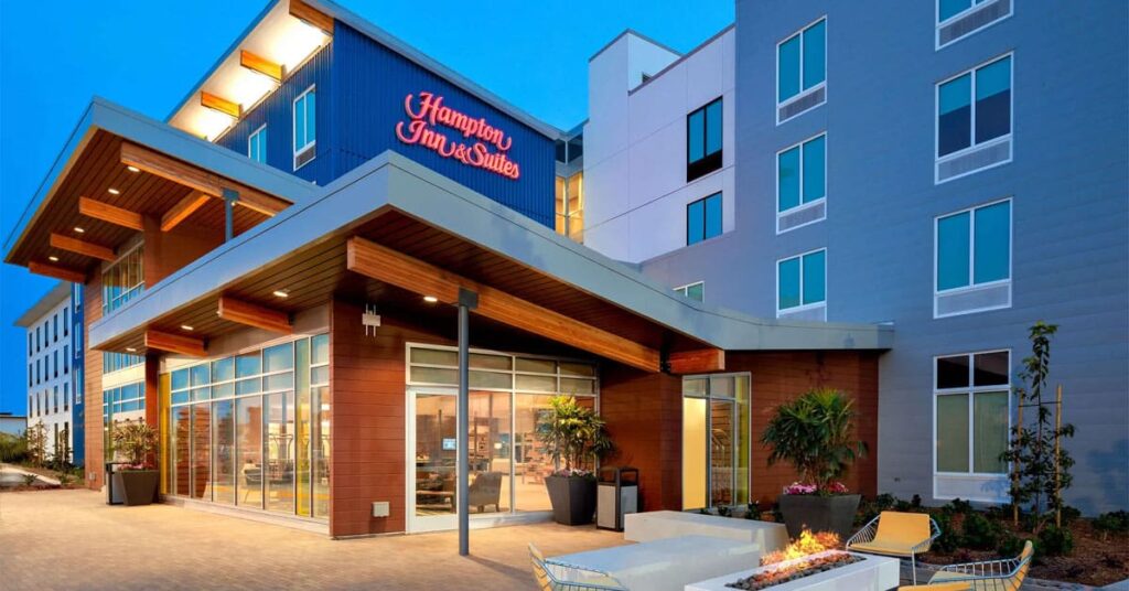 Modern Hampton Inn & Suites hotel exterior at dusk, featuring large windows, wooden and glass facade, illuminated sign, and an inviting outdoor seating area with a fire pit—perfect for Deeper Learning 2026 education events or relaxing by the entrance plants.