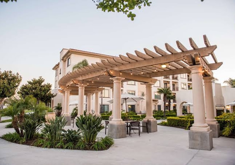 A beige pergola with columns stands in a landscaped courtyard featuring lush greenery and outdoor seating, creating an inspiring space for deeper learning in front of a multi-story building under a clear sky.