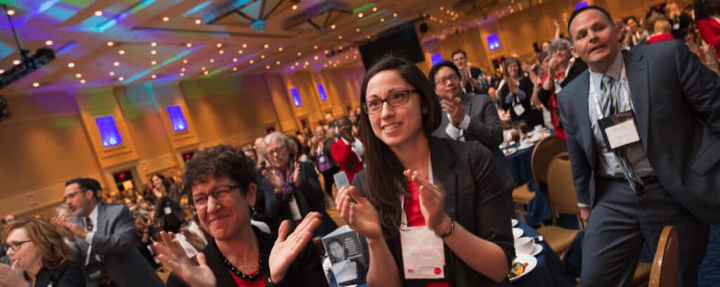 A group of people in business attire stand and enthusiastically applaud at the Summer Symposium in a large, well-lit room.