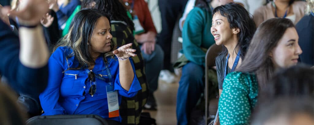 Two women sit facing each other in a crowded room at the Summer Symposium, engaged in an animated conversation. One gestures with her hand while the other smiles, surrounded by other Grade 9 students.