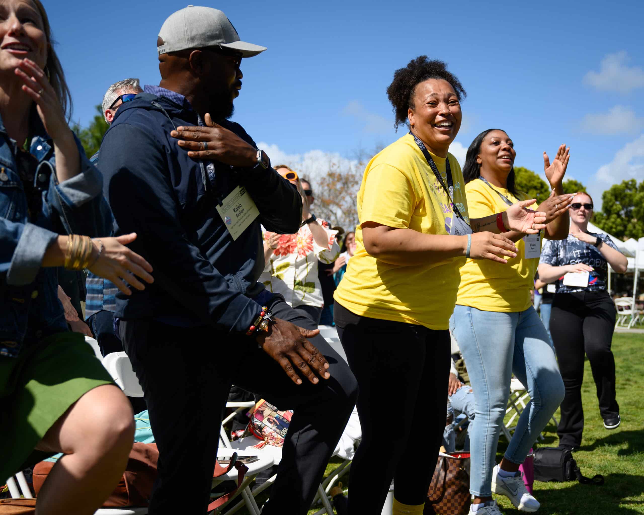A group of people, some wearing yellow shirts, stand and cheer outdoors on a sunny day. Smiling and clapping near white folding chairs, they enjoy a lively Deeper Learning 2026 event with blue sky and green grass in the background.