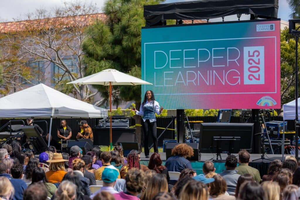 A speaker stands on an outdoor stage in front of a large screen that reads “Deeper Learning 2026,” addressing a seated audience at a daytime event. Trees and equipment surround the stage.