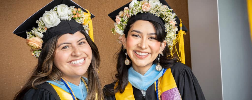 Two young women in graduation caps decorated with flowers and gowns smile happily at the camera, celebrating their achievement at the San Diego Teacher Residency. Join our Sep 2025 Information Session to learn how you can be next!.