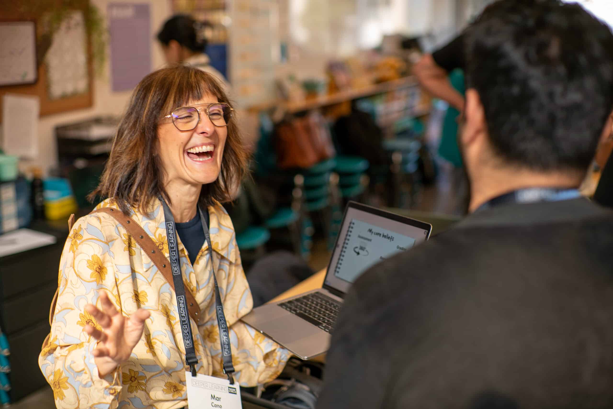 A woman with glasses and a floral shirt smiles and gestures while talking to a man. She holds a laptop displaying Deeper Learning 2026, and both wear name badges in a bright, busy education-focused indoor setting.