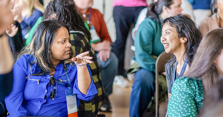 Two women sit facing each other in a lively group setting, engaging in animated conversation. One gestures while speaking, and the other woman listens with a smile. Blurred people are visible in the background.