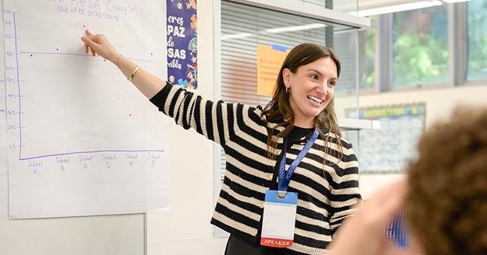 A woman wearing a striped sweater and conference badge smiles while pointing at a graph on a large paper chart during a presentation in a classroom.