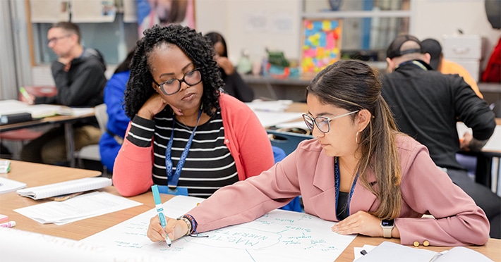 Two women sit at a table working together. One writes on a large sheet of paper with a marker while the other looks on. Papers, notebooks, and other people are visible in the background.