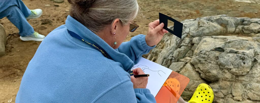 A Youth Changemakers participant in a blue fleece and glasses sketches on a clipboard outdoors, holding up a small black rectangular tool. Rocks and another person’s legs and shoes are visible in the background, showcasing Solutionary PBL in action.