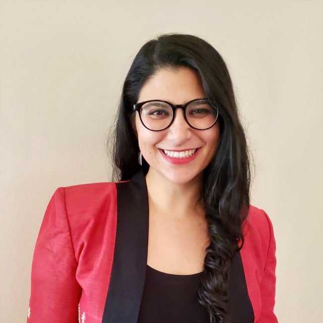 Gitanjali Paul, a woman with long dark hair and glasses, smiles at the camera. She is wearing a red blazer with a black top underneath, standing against a plain beige background.