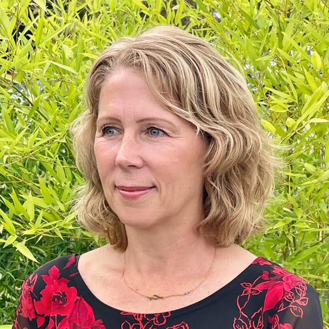 Kacey Dewing, a woman with wavy blonde hair wearing a black top with red floral patterns, stands in front of green leafy plants, looking slightly to the side and smiling softly.