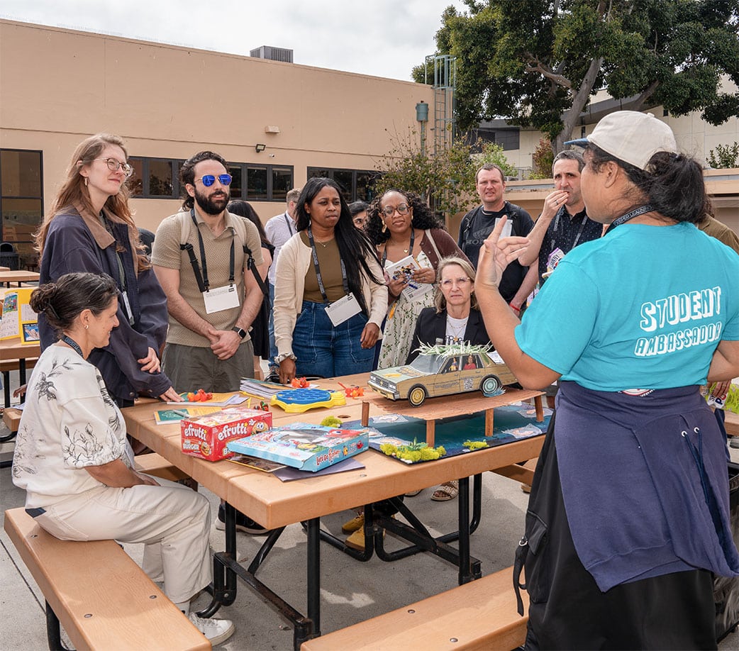 A group of adults gathers around a table at the BIG Event, listening to a Student Ambassador explain model projects and games using an event template. The setting appears to be a school or community gathering outdoors.