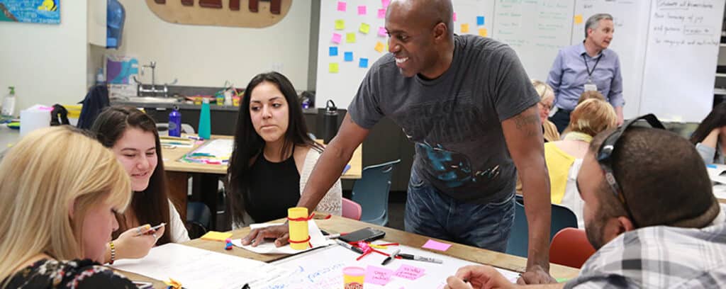 A group of adults collaborates at a classroom table covered with papers and Post-it notes, tackling real problems together. A man stands and smiles, engaging with the group as a teacher talks in the background, fostering continuous improvement.