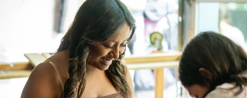 A woman with long hair smiles while looking down, sitting next to another person indoors near a window with bright natural light and eco-friendly design elements.