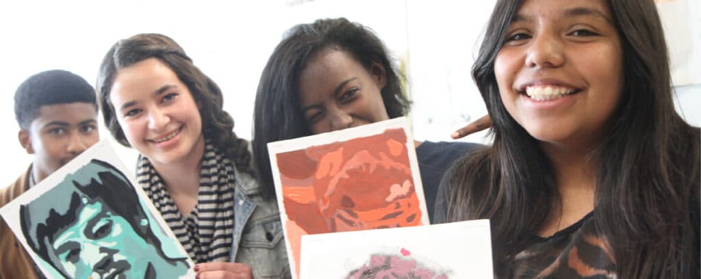 Four smiling teenagers hold up colorful portrait artwork, standing closely together. Their expressive creations reflect strong foundations in art and a spirit of continuous improvement as they celebrate their creative achievements.