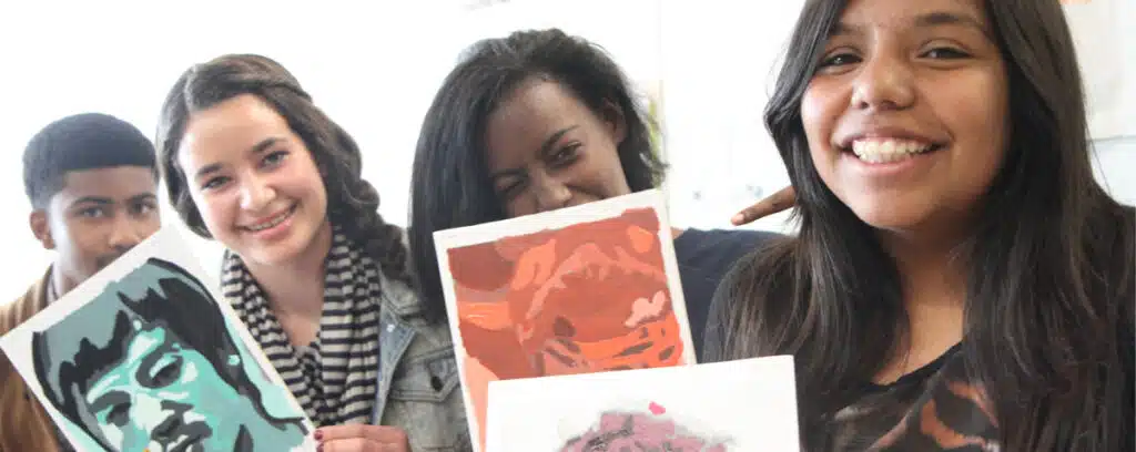 Four smiling teenagers hold up colorful portrait artwork, standing closely together. Their expressive creations reflect strong foundations in art and a spirit of continuous improvement as they celebrate their creative achievements.