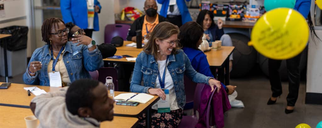 Adults sit at tables in a classroom at the East Coast Summit, some smiling and interacting with a yellow balloon floating in the air. Notebooks, coffee cups, and snacks are on tables as people engage in conversations about education and improvement.