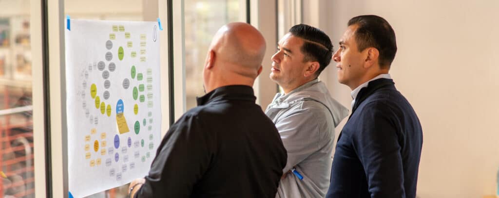 Three men stand indoors, examining a large process mapping chart taped to a window. The colorful diagram of interconnected circles and labels sparks a focused discussion on how to improve outcomes.
