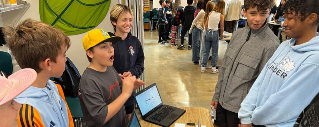 A group of students stand around a table with a laptop, playfully debating as they play it by ear during their Project Unpacking. More students fill the background, adding to the lively classroom atmosphere.