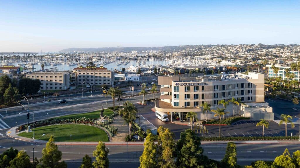 Aerial view of a DoubleTree hotel near a marina with numerous boats docked, perfect for your next BIG Event. The scene features palm trees, streets, and buildings, with a coastal city and hills in the background.