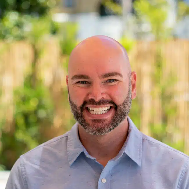 A smiling bald man with a trimmed beard wearing a light blue collared shirt stands outdoors with a blurred background of greenery and a wooden fence.