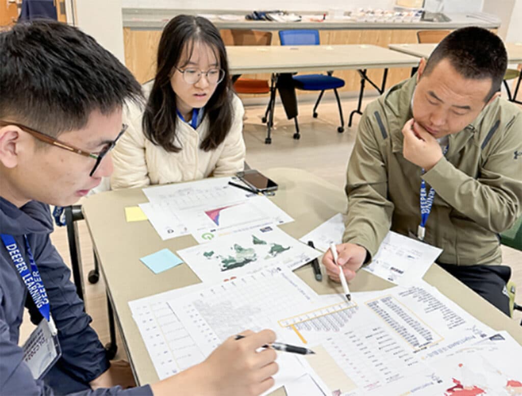 Three people sit at a table, closely examining and discussing various printed charts, graphs, and documents spread out in front of them in a well-lit room, focused on advancing Deeper Learning within global education.