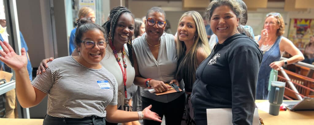 Five smiling women stand close together indoors, some with arms around each other at an Educational Leadership information session. One woman in front wears a striped shirt and glasses, holding her hands up in joy, while others hold clipboards or papers.
