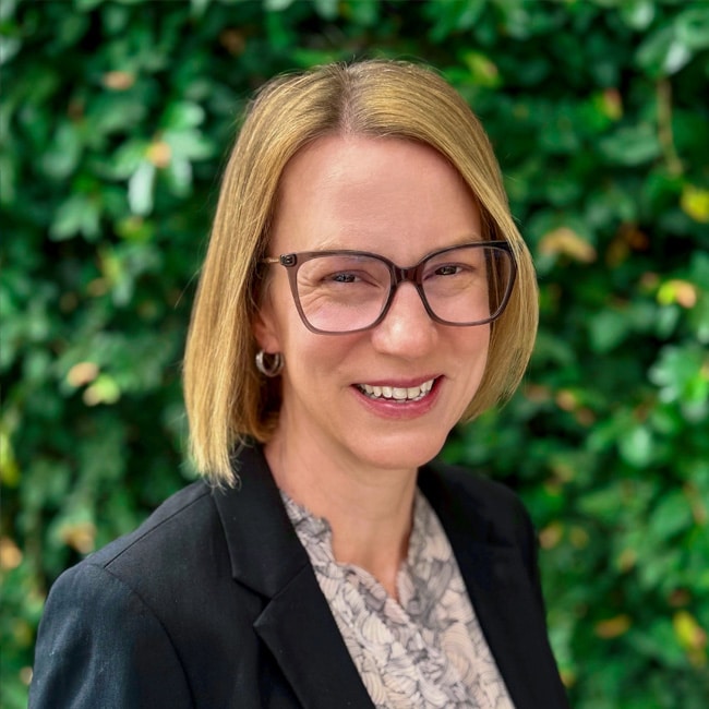 Sara Gallagher, a woman with straight, shoulder-length blonde hair and glasses, smiles at the camera. She is wearing a black blazer and light-patterned blouse, standing in front of a leafy green background.