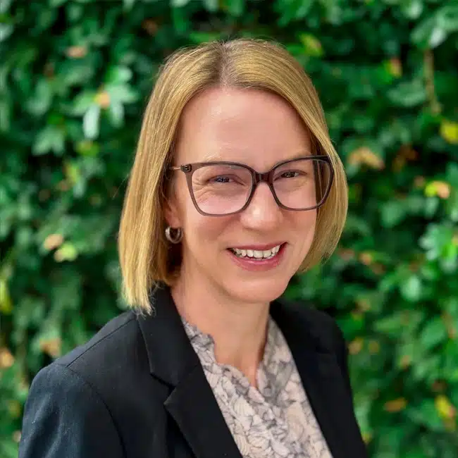 Sara Gallagher, a woman with straight, shoulder-length blonde hair and glasses, smiles at the camera. She is wearing a black blazer and light-patterned blouse, standing in front of a leafy green background.