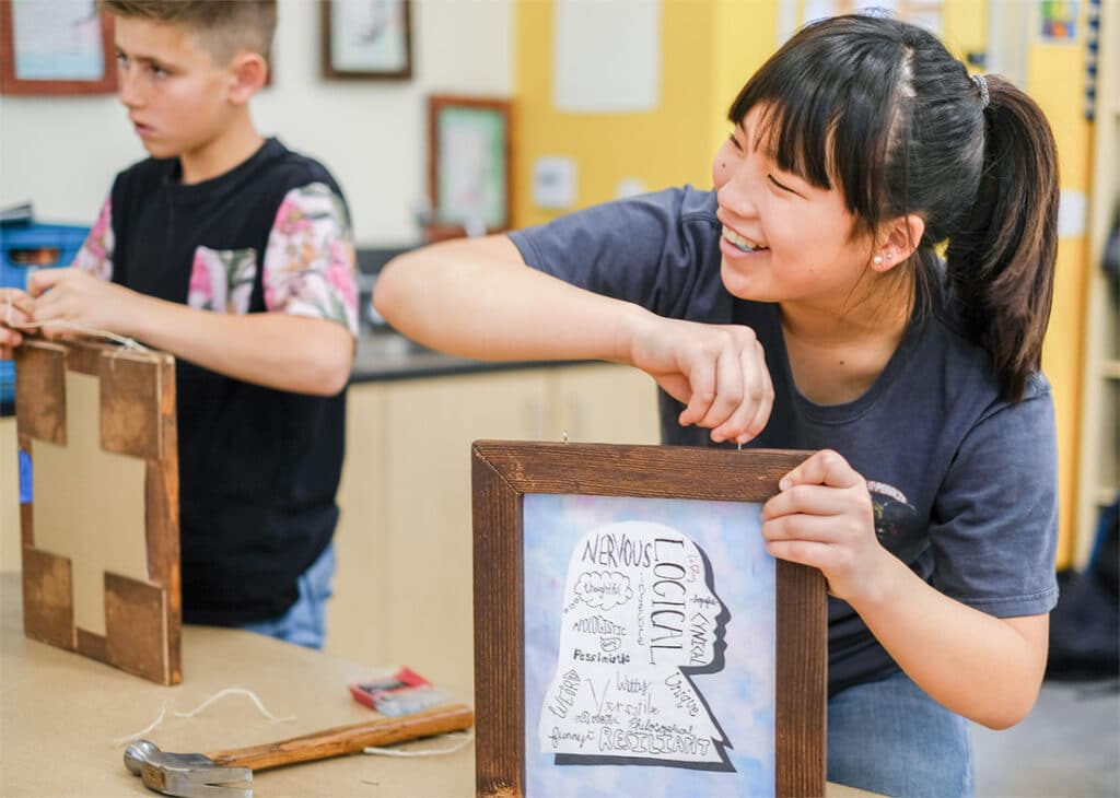 A smiling girl attaches a wooden frame to a paper with handwritten words, using Unboxed materials, while a boy beside her works on a similar project at a classroom table scattered with creative tools.