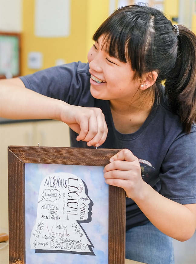 A smiling young woman with a ponytail holds an Unboxed framed artwork showing a silhouette filled with handwritten words like loyalty, nervous, resilient, and passionate in various fonts.