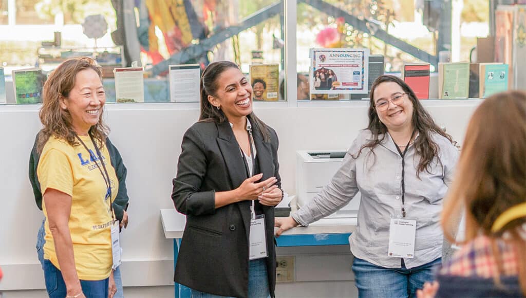 Three women stand together indoors, smiling and laughing during a casual group conversation. Name badges hang from their necks, and books are displayed on a shelf behind them. Sunlight shines through large windows.