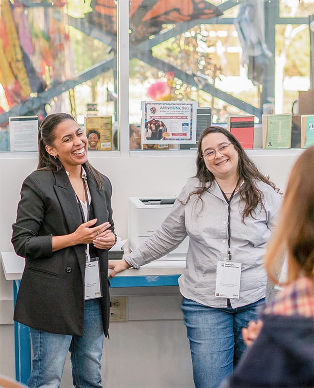 Two women wearing conference badges stand and smile in front of a small group indoors. One is wearing a black blazer and the other a gray shirt. Colorful decorations and flyers are visible in the background.