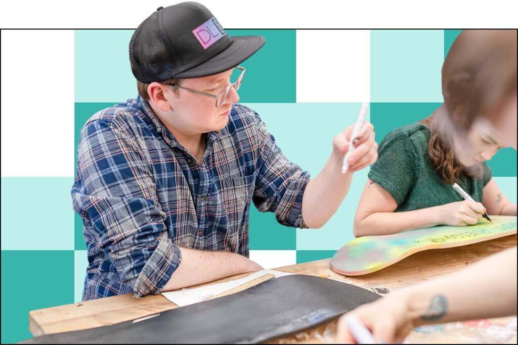 A man in a plaid shirt and cap writes on paper at a table, engaging in deeper learning, while a woman next to him decorates a skateboard. The background features teal and white checkered squares.