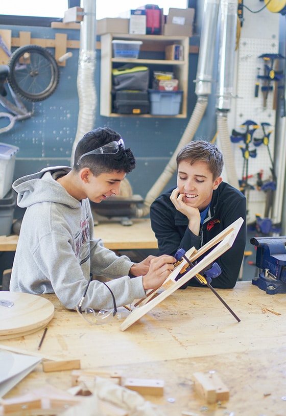 Two young people smile and work together on a woodworking project in a workshop, experiencing deeper learning as they use tools at a large workbench surrounded by shelves, equipment, and supplies.