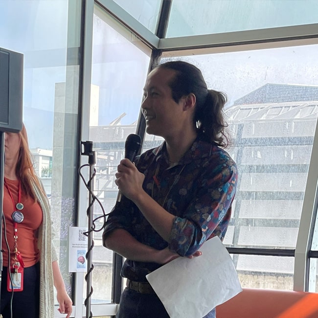 A person with long hair in a ponytail holds a microphone and a paper, smiling while speaking indoors near large windows. Another person stands nearby, partially out of view.