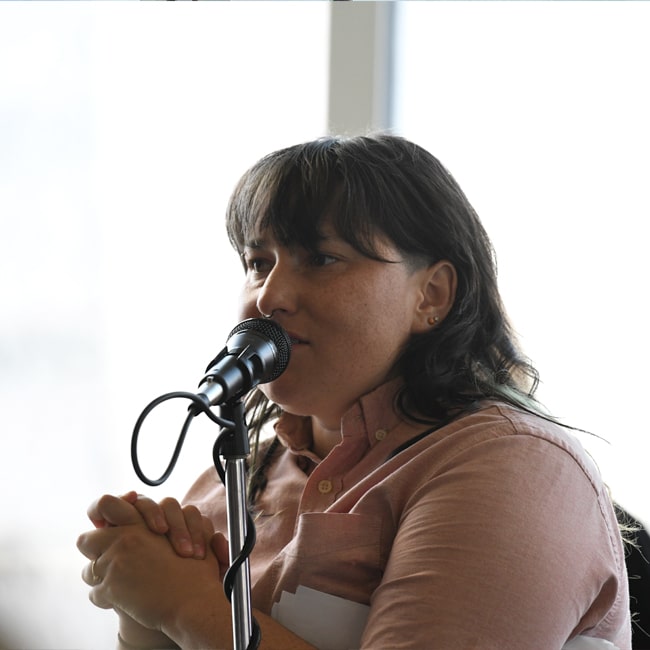 A person with shoulder-length dark hair speaks into a microphone, sitting indoors with hands clasped and wearing a light pink shirt. The background is softly blurred.