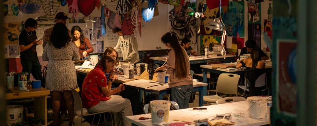 A group of people work on art projects at tables in a colorful, busy High Tech High art studio filled with paintings, drawings, and supplies. The room is warmly lit, and everyone is focused on their creative activities and Innovation Residency projects.