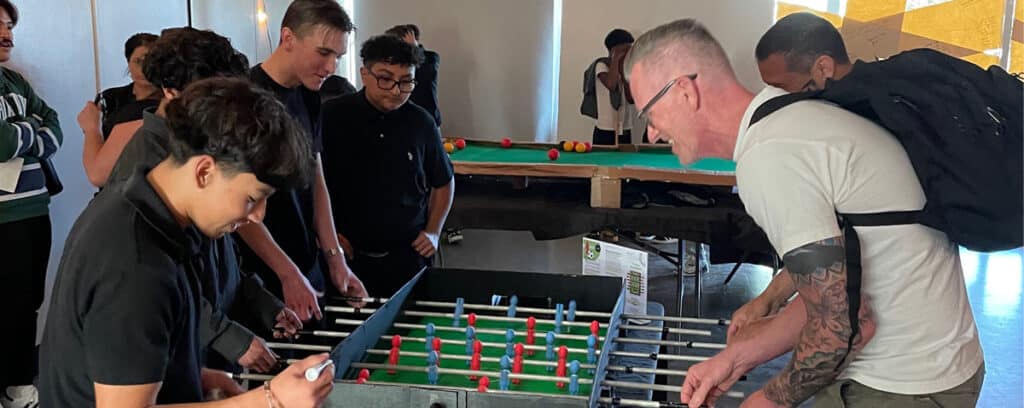 A group of people enthusiastically play foosball indoors during a Project Unpacking event, with another group enjoying a similar tabletop game in the background. The atmosphere appears lively and social.