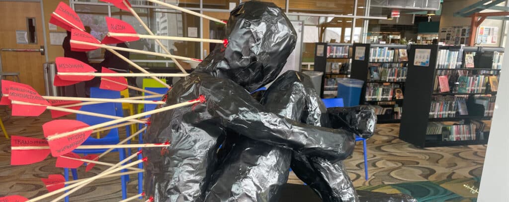 A black human figure sculpture, part of Project Unpacking, sits with knees drawn up, pierced by arrows with red fletching labeled “racism,” “sexism,” and “homophobia,” in a library setting with bookshelves in the background.