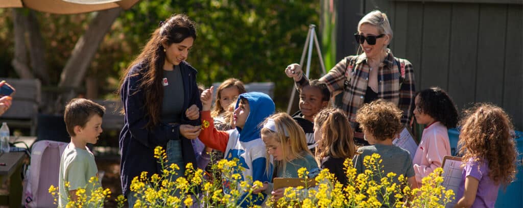 Two adults and a group of young children stand outside in a garden with yellow flowers. The children appear engaged and curious, some holding clipboards, as the San Diego Teacher Residency (SDTR) team interacts and guides them.