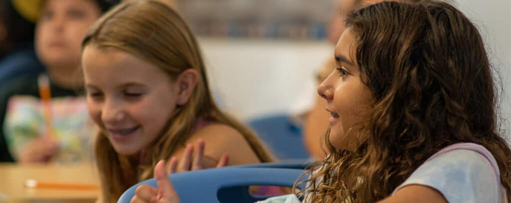 Two young girls sit in a classroom, smiling and talking with each other. They appear engaged and happy, sharing their student voices as blurred classmates work in the background.