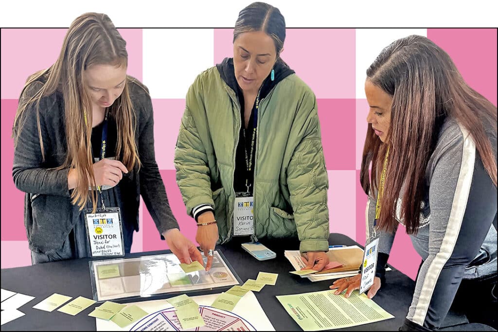 Three women with name badges stand around a table, closely examining documents and colored paper cards at what appears to be a workshop or educational event. Pink and white checkerboard background.