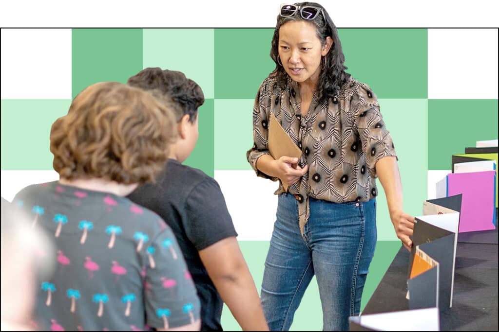 A woman in a patterned blouse and jeans talks to two children standing by a table displaying colorful folders or boxes, with a green and white checkered background.